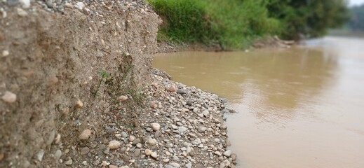Eroding riverbank with visible layers of soil and rocks next to a muddy river, lush green vegetation on the opposite bank.