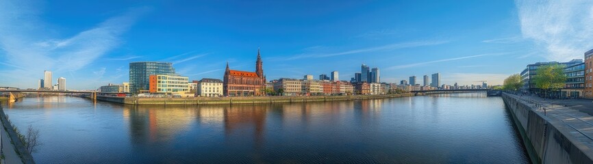 Obraz premium Panoramic view of a city skyline reflecting on a calm river. Modern buildings and a historical church line the waterfront on a clear, sunny day