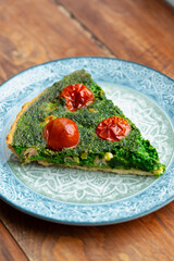Delicious slice of spinach and tomato quiche served on decorative blue plate on wooden table