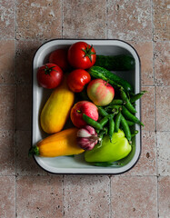 Fresh garden organic vegetables and fruits in an enameled tray on a stone background, top view