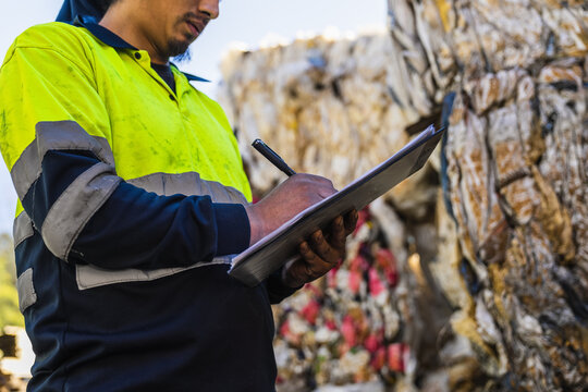 Waste management worker writing notes on clipboard while inspecting a large pile of recycled materials at a recycling center - Powered by Adobe