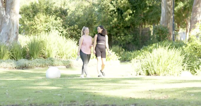 Diverse female friends walking past sculpture in park stopping, chatting, pointing and laughing