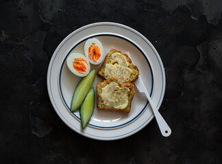 Delicious breakfast - toasts with butter, boiled egg, fresh cucumber on a plate on a dark background, top view