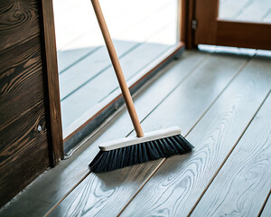 Wooden-handled black broom resting on a painted gray wooden deck near an open door. Escoba tradicional de mango de madera y cerdas negras sobre una terraza de madera gris junto a una puerta abierta.

