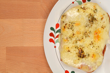 An appetizing toasted sandwich with melted cheese and herbs rests on a decorative plate, set against a warm wooden background, shot from above