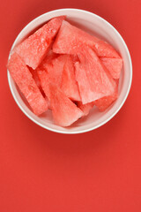 A white bowl brimming with bright red watermelon pieces  on a vibrant red background, offering a refreshing view