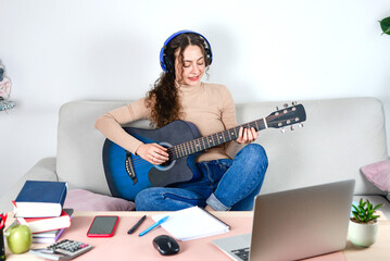 A young woman is enjoying playing her guitar at home while using her laptop with headphones on