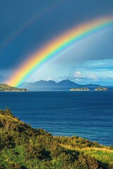 Majestic rainbow arches over a tranquil ocean scene. Lush green hills meet the deep blue water, with distant islands framed by the vibrant arc of the rainbow