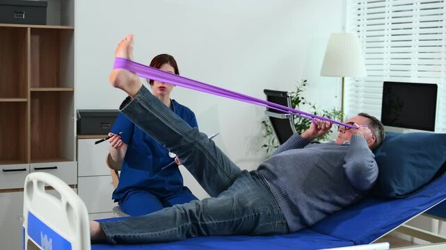 Senior man exercises with resistance band in bed during home rehab, supervised by nurse. Concept of recovery, strength, physiotherapy, and elderly care. Real people scene.