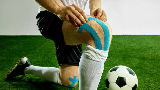 Soccer player applying blue kinesiology tape on knee on green field. Concept of sports recovery, football injury prevention, kinesiology taping, male athlete health preparation.