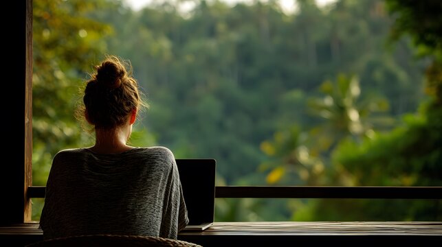 Back view of female remote worker seated on a deck laptop in front off frame lush jungle visible in background natural textures and relaxed digital nomad life