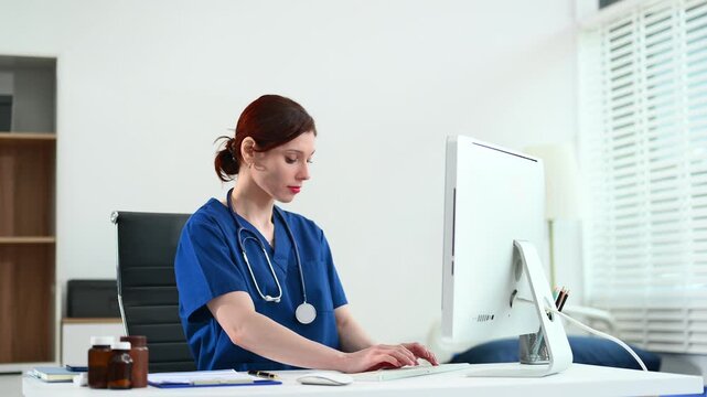Female doctor in scrubs works at a desktop computer in bright office. Concept of modern healthcare, digital records, and admin tasks. Real people, clinical setting.