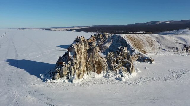 Aerial view of jagged rocks rising from the frozen expanse of Lake Baikal, where the stark white ice meets the rugged, snow-dusted terrain, Irkutsk Oblast, Russia.