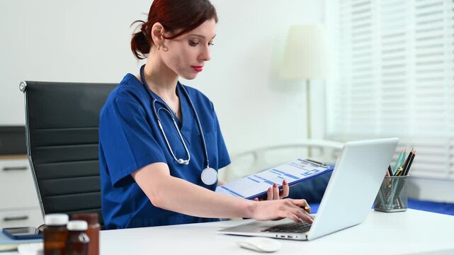Female doctor in scrubs works at a desktop computer in bright office. Concept of modern healthcare, digital records, and admin tasks. Real people, clinical setting.