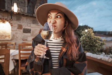 Funny young woman in hat grimacing while smelling white wine in glass, outdoor restaurant setting, evening ambiance