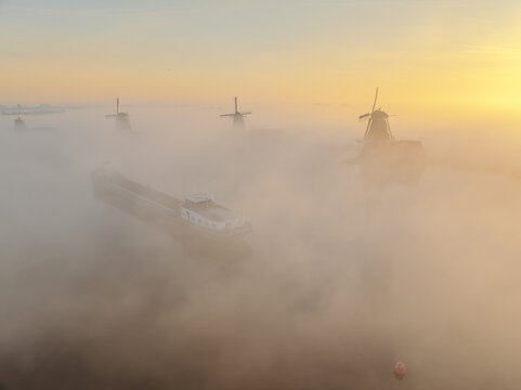 Aerial view of windmills and a barge veiled in a thick fog at dawn, creating an ethereal landscape, Zaandam, Noord-Holland, Netherlands.
