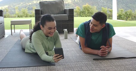 Spotting funny clip, diverse couple on covered patio scrolling smartphone and laughing on yoga mats - Powered by Adobe