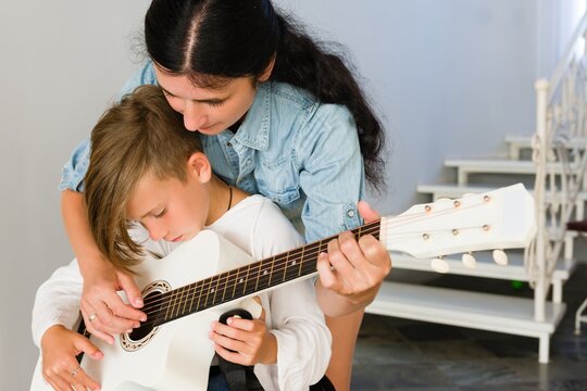 A woman patiently teaches a boy to play the guitar. She guides his hands, fostering a loving and supportive learning environment.