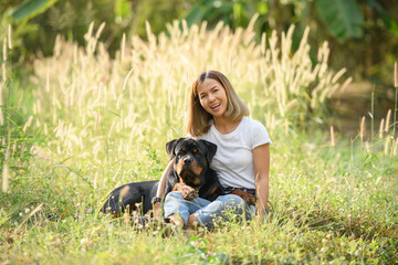 Happy Asian woman sitting in a field with her Rottweiler dog, enjoying nature and sunshine. Concept of pet love, outdoor lifestyle, and peaceful countryside moments.