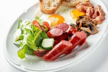 Hearty breakfast with sausages, bacon, fried eggs, mushrooms, salad, and bread slices served on a white plate. Colorful, fresh, and appetizing morning meal.