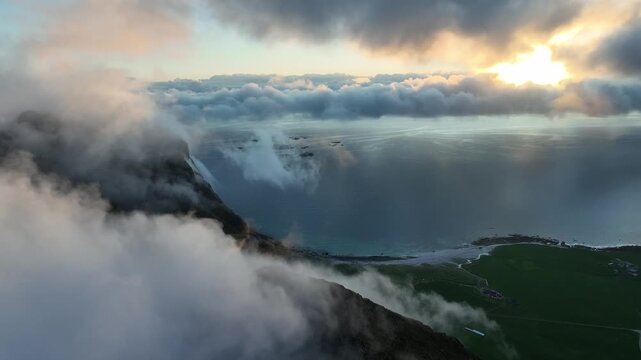 Drone ascends above foggy Mannen peaks with Utakleiv beach and ocean glowing in midnight sun