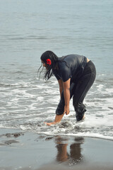 A teen girl, soaked from head to toe, bends forward as sea foam rushes past, red blossom tucked in...