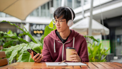 Glasses student man with headphone holding coffee looking at phone sits with notebook at cafe table.
