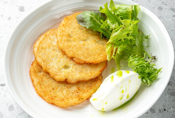 Crispy golden potato pancakes served with fresh mixed greens and sour cream, presented on a white plate over a light speckled background.
