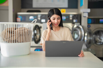 Smiling lady combining work and chores, talking on the phone and typing on a laptop with laundry in the background.