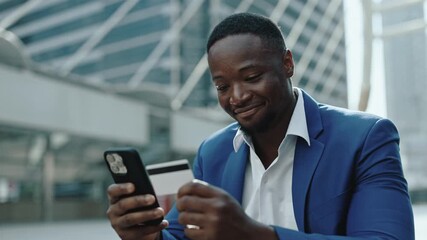 Portrait of happy African American businessman uses banking app on smartphone in urban district. Young entrepreneur pays with internet technology sitting on parapet near skyscraper - Powered by Adobe