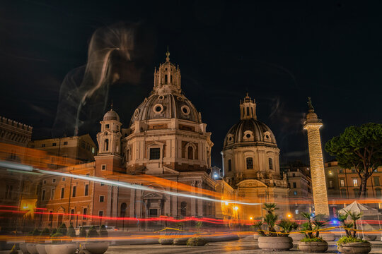 View of illuminated Trajan's Column and the domed churches against the dark sky, with streaks of light painting the bustling Roman night, Rome, Lazio, Italy.