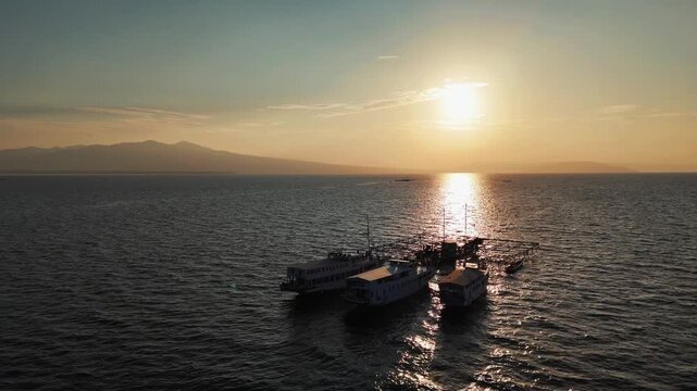 Aerial view of boats floating on dark ocean waters as the sun sets, casting a golden glow across the scene, Poto Tano, Indonesia.
