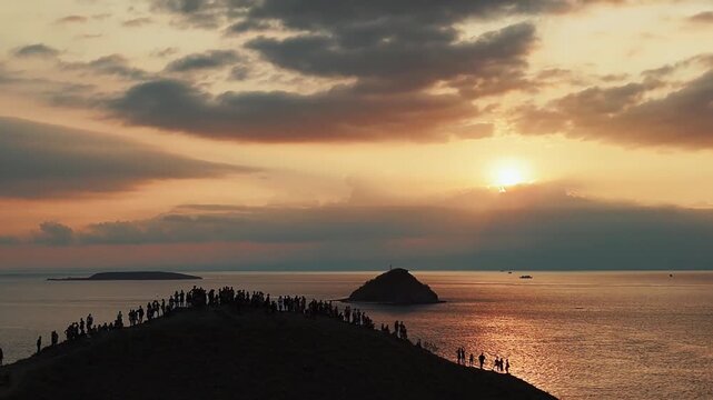 Aerial view of people silhouetted against the setting sun, gathering on a hilltop overlooking the ocean at dusk, Poto Tano, Indonesia.