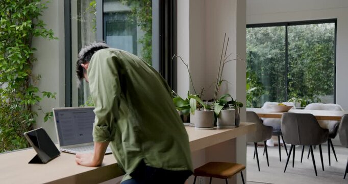 Man is entering kitchen due to deadline, sitting on stool typing on laptop then standing