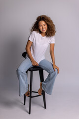 Curly Woman Sitting On Stool In White T-Shirt And Blue Jeans