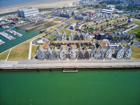 Aerial view of modern, geometric buildings lining the waterfront, contrasting with the bright green water and distant sandy beach, Dunkirk, France.