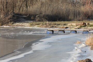 Frozen lake with wooden pier on blue barrel in ice in early springtime