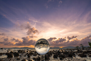  A mesmerizing sunset over a rocky shoreline captured through a glass crystal ball, creating a surreal inverted reflection of the seascape beneath vibrant, colorful skies.