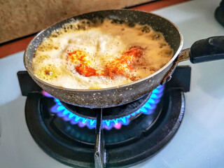 Close-up of an egg frying in bubbling oil in a small teflon pan over a blue gas flame on a stove, capturing home cooking with vivid colors and sizzle detail.
