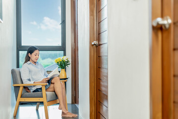 A young Asian woman reads a book on a couch on her porch at home on a weekend without electronics. The concept of digital detox and social media. Reading is a leisure activity.