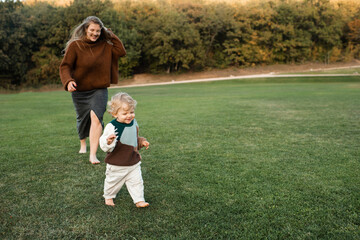 Smiling woman running with baby girl 1-2 year old on green grass lawn outdoors. Motherhood.