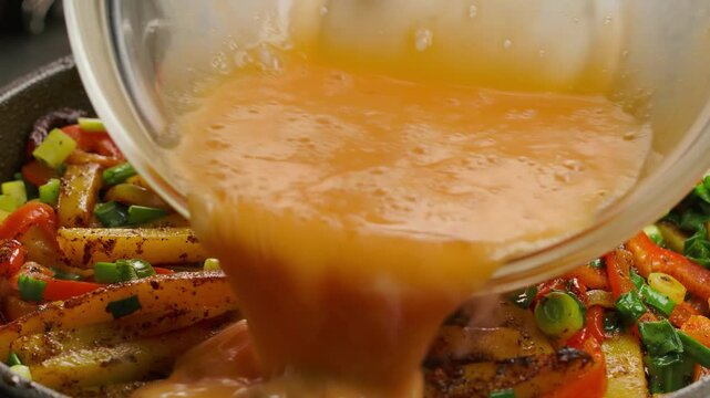 Cooking a hearty breakfast. Whisked eggs are poured over a colorful mix of sizzling potatoes, peppers, and onions in a hot skillet, creating a delicious scramble or frittata closeup