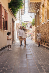 A family walks together in a narrow street. The mother and father, both young adults, are smiling. Their son and daughter play nearby, enjoying the sunny day.