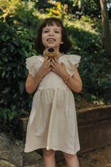 A young girl with brown hair smiles while holding a stuffed animal. She wears a light-colored dress and stands in a lush green garden.