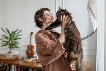 Happy loving young woman holds fluffy cat smiling, making playful kissy face spending pleasure time...