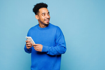 Young man of African American ethnicity wear sweatshirt casual clothes hold in hand use mobile cell phone look aside isolated on plain pastel light blue background studio portrait. Lifestyle concept.