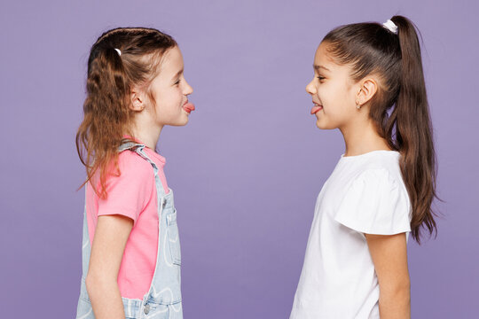 Side profile view smiling fun little two friends kids children girls 9-10 years old wear casual clothes together look to each other show tongue isolated on plain purple background. Lifestyle concept.