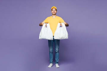 Full length young delivery guy employee man wear yellow cap t-shirt uniform workwear work as dealer courier hold in hand two plastic package bags isolated on plain purple background. Service concept.