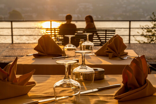 Restaurant tables set for dinner and couple overlooking lake at sunset, Como, Province of Como, Lake Como, Lombardy, Italian Lakes, Italy