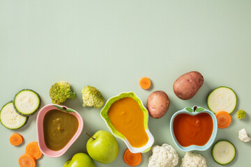 Three apple-shaped bowls with colorful vegetable purees - green, orange, and red, surrounded by fresh ingredients: zucchini, carrot, potato, broccoli, cauliflower, and green apple